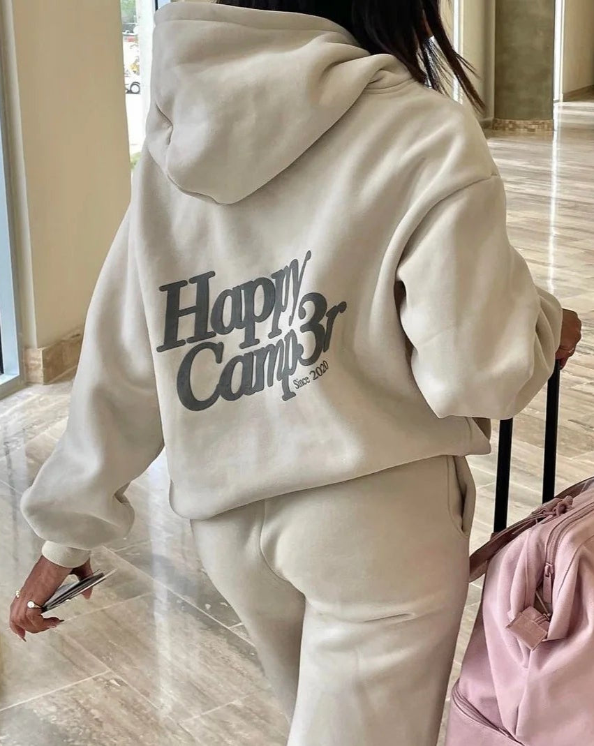 Person wearing a 'Happy Camper' hoodie with a pink suitcase in a lobby.