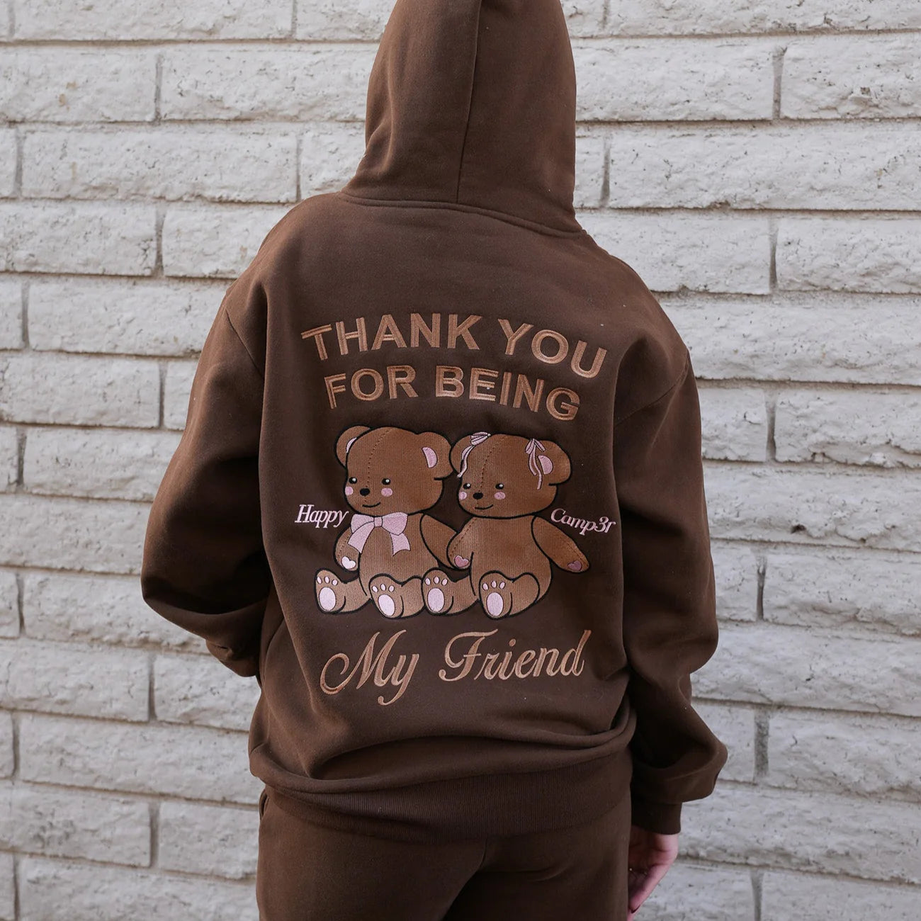 Brown hoodie with teddy bears and text on the back against a brick wall.