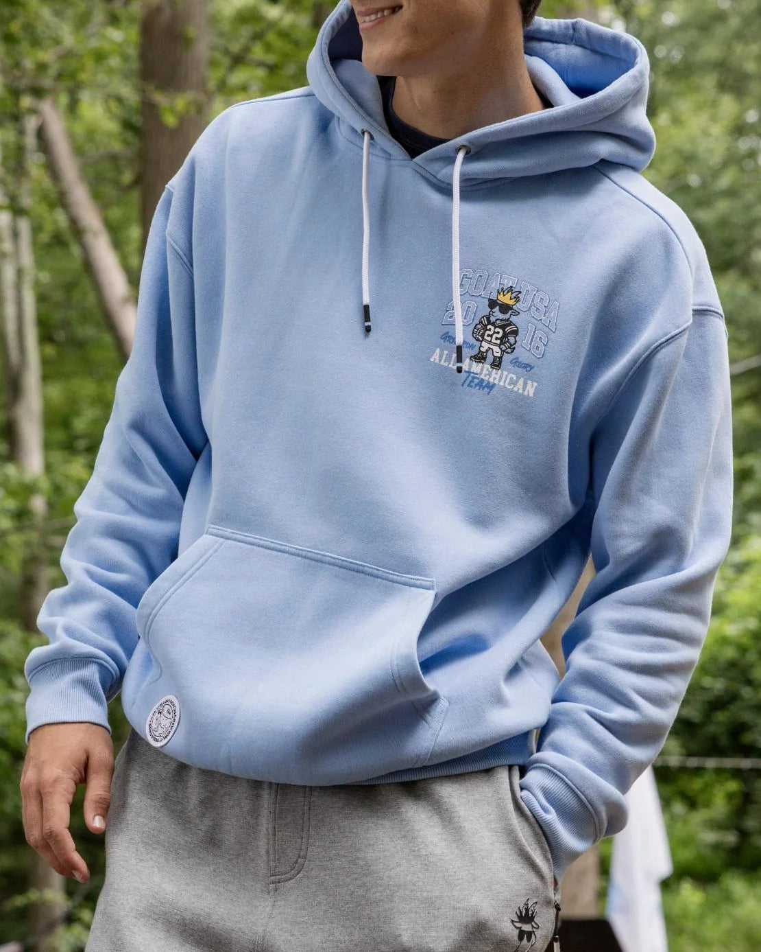 Person wearing a light blue hoodie with a logo outdoors in a forest setting