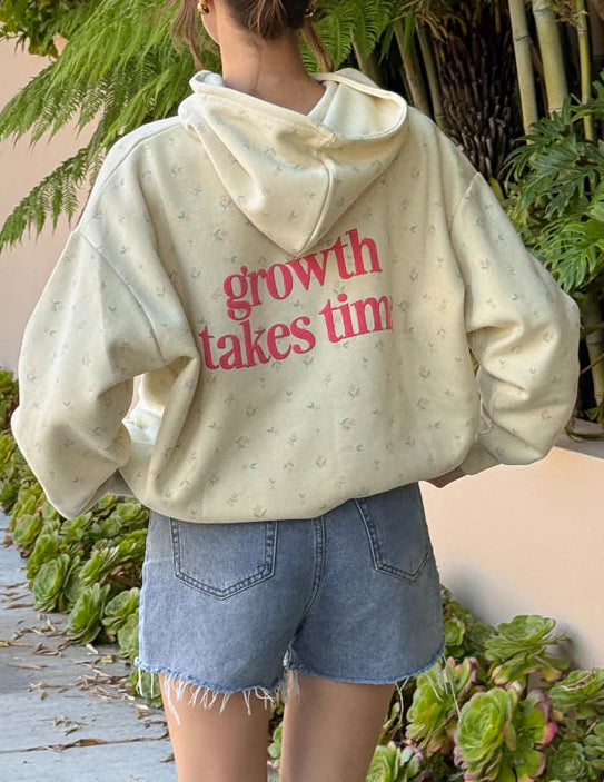 Person wearing a hoodie with 'growth takes time' text, standing outdoors near greenery.
