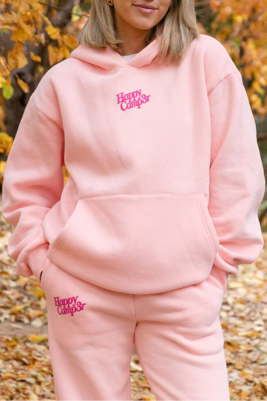 Person wearing a pink 'Happy Campers' hoodie and pants in an autumn setting.