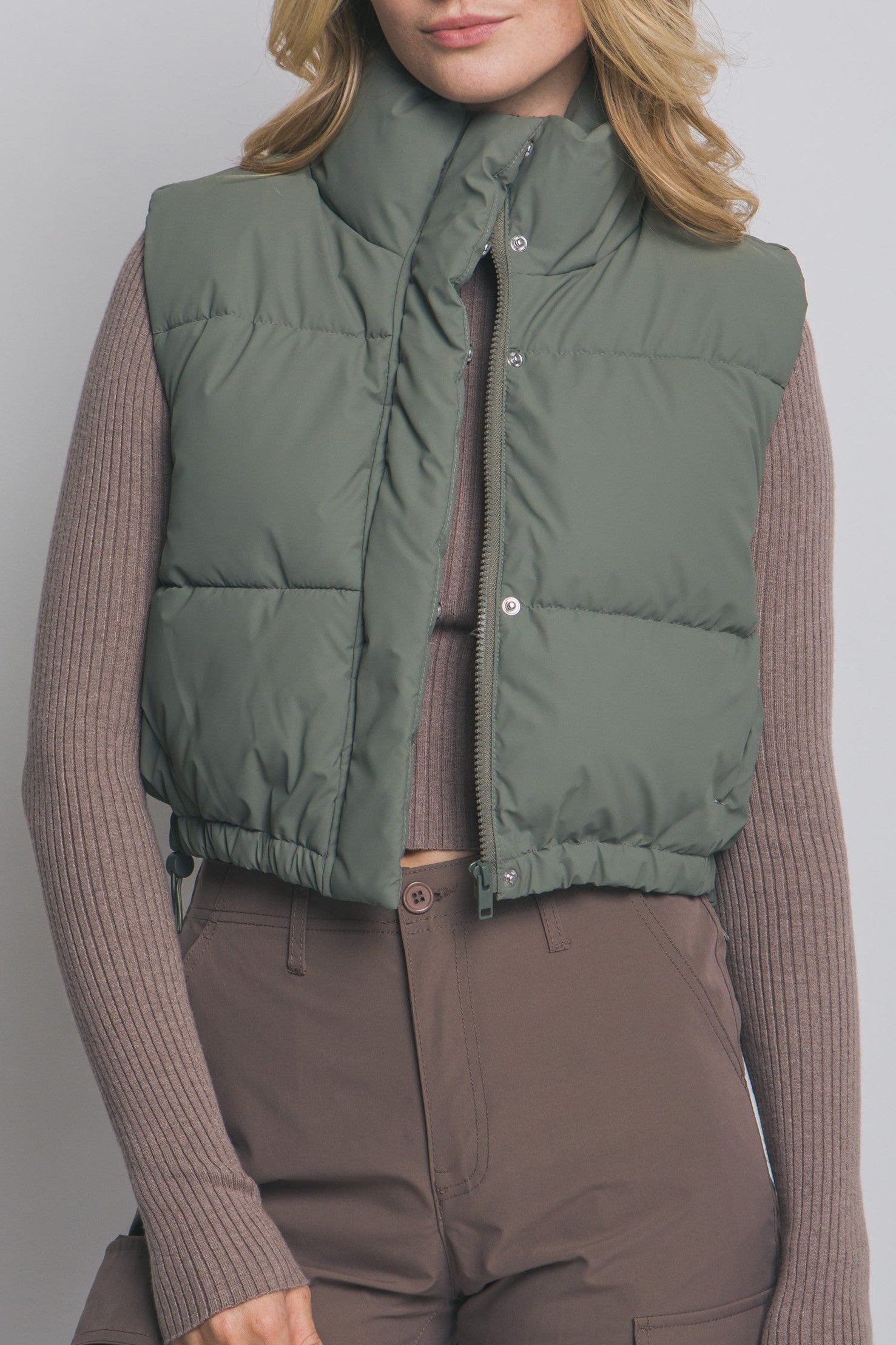 Woman wearing a green puffer vest over a brown sweater and pants on a plain background