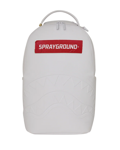 A white Sprayground backpack featuring a red rectangular logo patch on the front with white text, a top carry handle, and an embossed shark-face pattern on the lower half, shown against a plain white background.