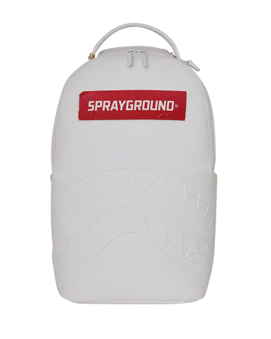 A white Sprayground backpack featuring a red rectangular logo patch on the front with white text, a top carry handle, and an embossed shark-face pattern on the lower half, shown against a plain white background.