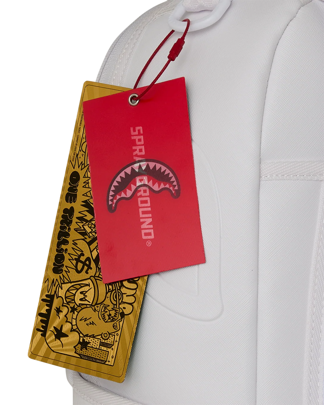 A close-up of a white garment with a red and yellow hanging tag. The red tag has the text 'SPRAYGROUND' and a shark-mouth graphic, while the yellow tag has black graffiti-style artwork and text.