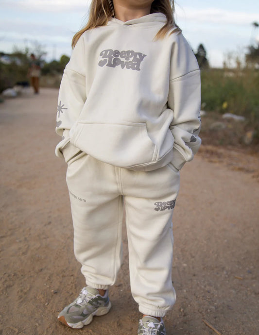 Person wearing a beige hoodie and pants with 'Deeply Loved' branding outdoors.
