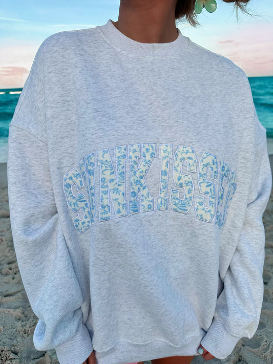 Person wearing a gray sweatshirt with a blue patterned text design on a beach.