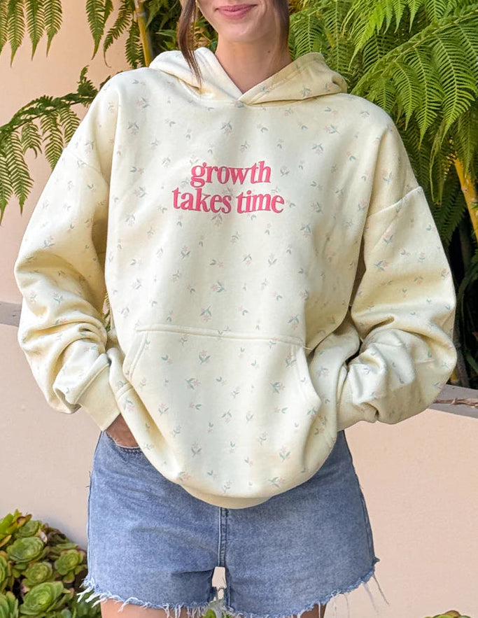 Person wearing a cream hoodie with 'growth takes time' text, standing outdoors with greenery. 