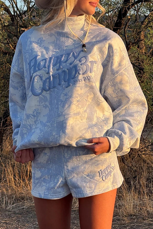 Person wearing a light blue 'Happy Camper' sweatshirt and shorts outdoors.