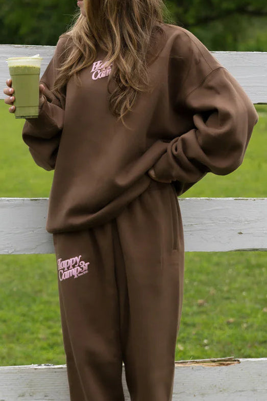 Person wearing a brown 'Happy Camper' tracksuit holding a green smoothie.