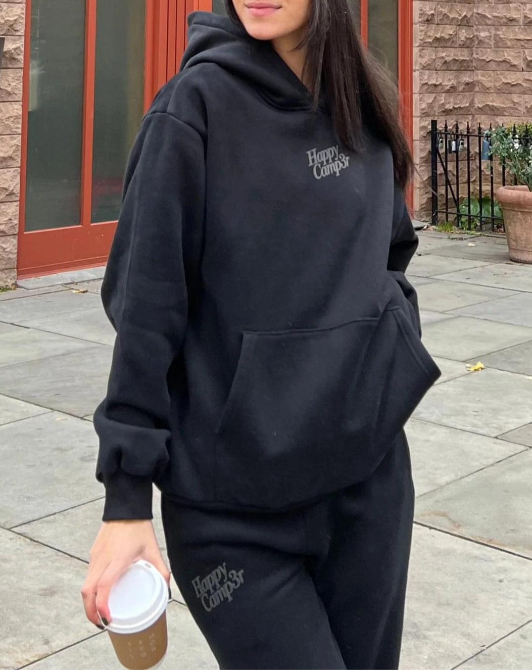 Person wearing a black hoodie and pants with text, holding a coffee cup, standing in front of a building.