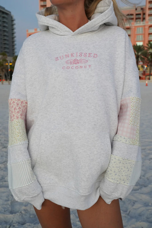 Person wearing a light gray hoodie with 'Sunkissed Coconut' text on a beach.