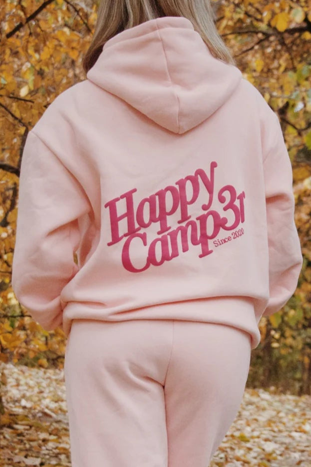 Person wearing a pink 'Happy Camper' hoodie and pants walking through a forest with autumn foliage. 