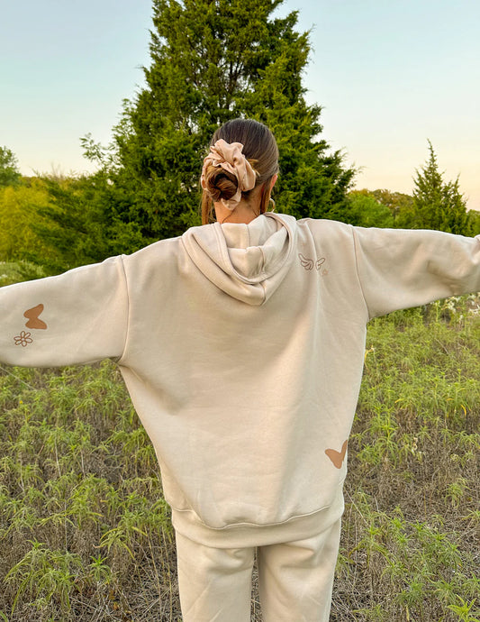 Person wearing a beige hoodie and pants standing in a field with trees in the background 