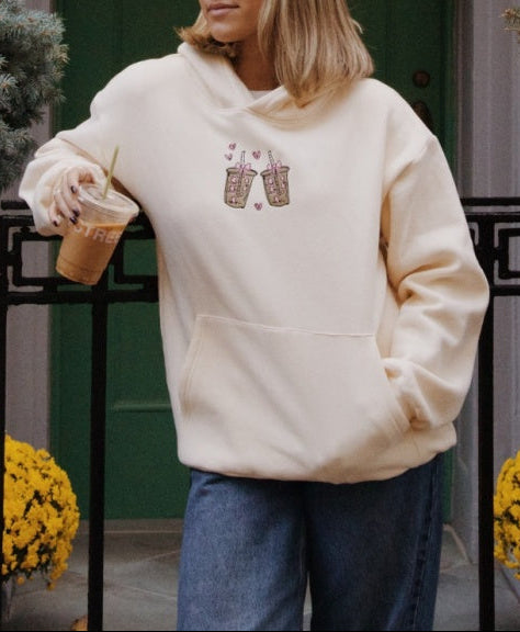 Person wearing a cream-colored hoodie with a graphic design, standing in front of a building with columns and greenery.